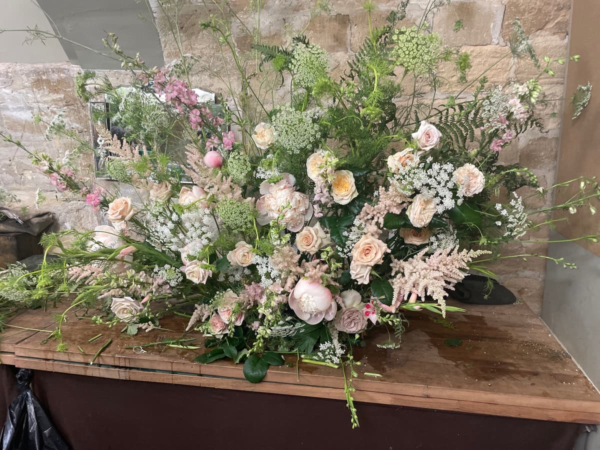 A large, sprawling floral arrangement on a wooden table against a stone wall, featuring blush pink peonies, peach garden roses, pink astilbe, and white Queen Anne's lace mixed with abundant greenery in a wild, garden-style design.