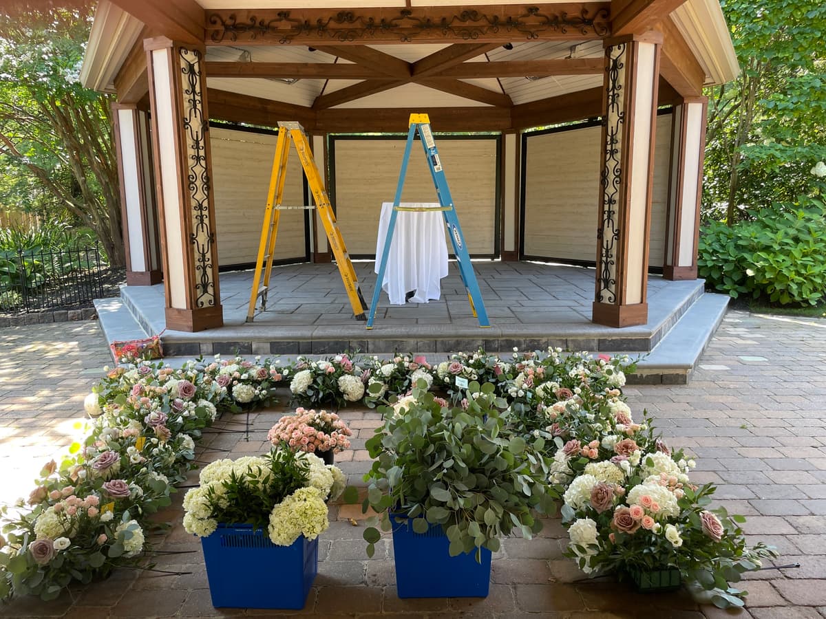 A behind-the-scenes look at a wedding floral setup featuring a semi-circle of lush flower arrangements in blue crates on a paved patio. The arrangements include white hydrangeas, dusty rose and blush roses, and eucalyptus. In the background, a wooden gazebo contains two ladders and a small table with a white tablecloth.