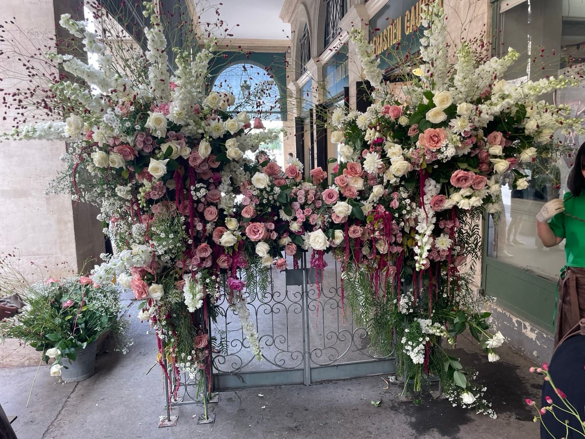A lavish floral installation covering a grey metal gate with cascading white delphiniums, pink and mauve roses, and hanging burgundy amaranthus tassels in an outdoor covered arcade.