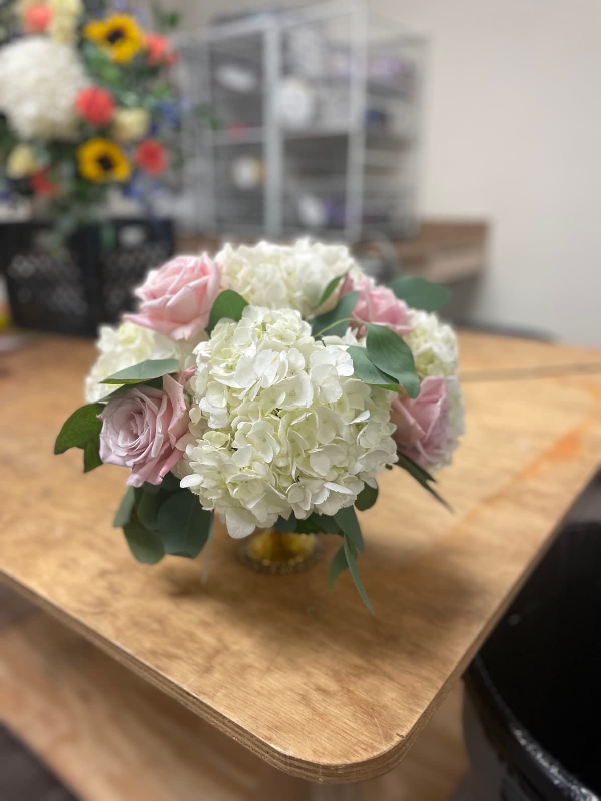 A lush, rounded floral centerpiece sitting on a wooden work table. The arrangement features large, fluffy white hydrangeas and soft blush pink roses nestled in a gold compote vase, accented with silver dollar eucalyptus leaves. The background shows a blurred floral studio setting.