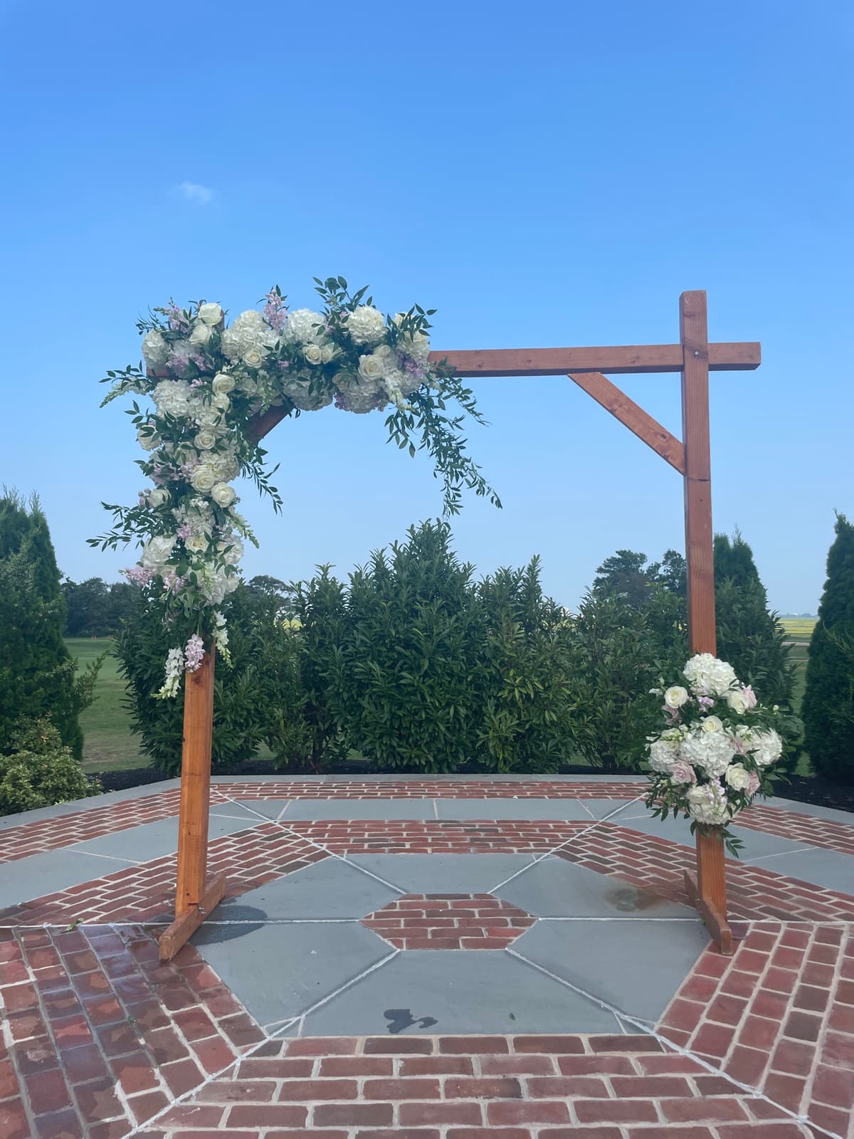 A wooden wedding arch stands on a brick and stone patio under a clear blue sky, decorated with asymmetrical floral arrangements featuring white roses, hydrangeas, and trailing greenery.