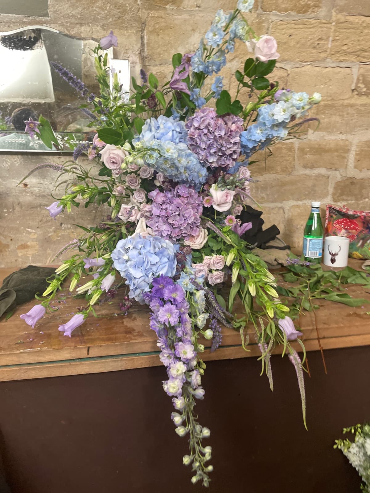 A lush, garden-style floral arrangement featuring blue hydrangeas, tall blue delphiniums, lavender roses, and purple bellflowers set against a rustic stone wall.