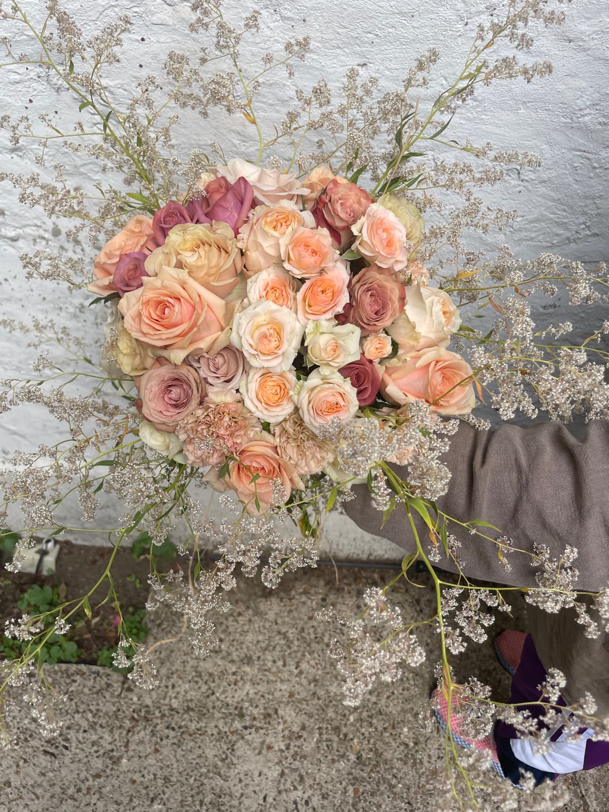 A top-down view of a lush, hand-tied bouquet held against a white textured wall. The arrangement features a dense cluster of roses in shades of peach, blush pink, cream, and dusty mauve, mixed with ruffled beige carnations. The central blooms are surrounded by a wide, airy halo of delicate white and silver filler flowers, creating an ethereal effect.