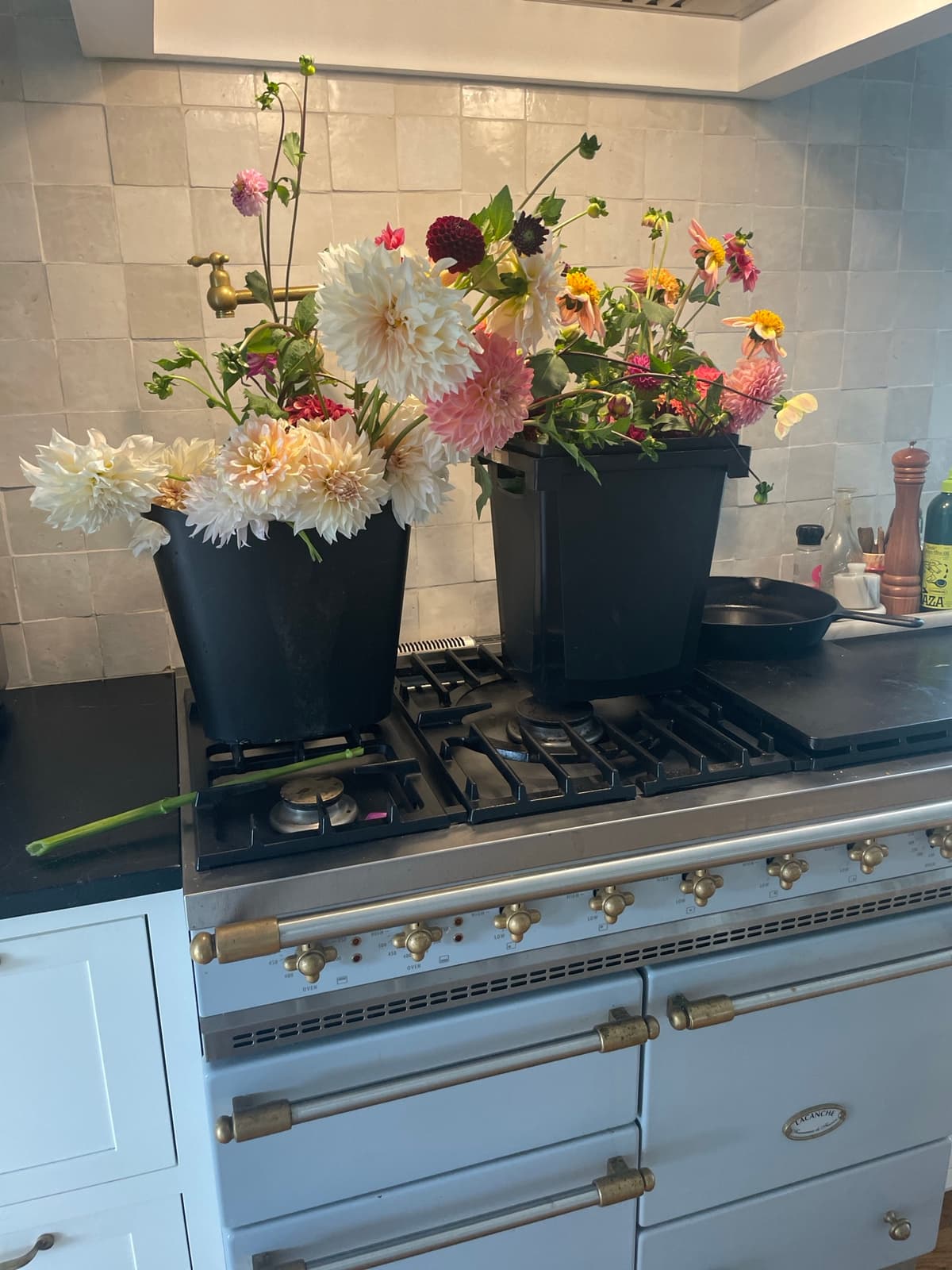 Two black buckets filled with freshly cut dahlia flowers in shades of white, pink, and burgundy sitting on a light blue Lacanche kitchen range against a tiled backsplash.