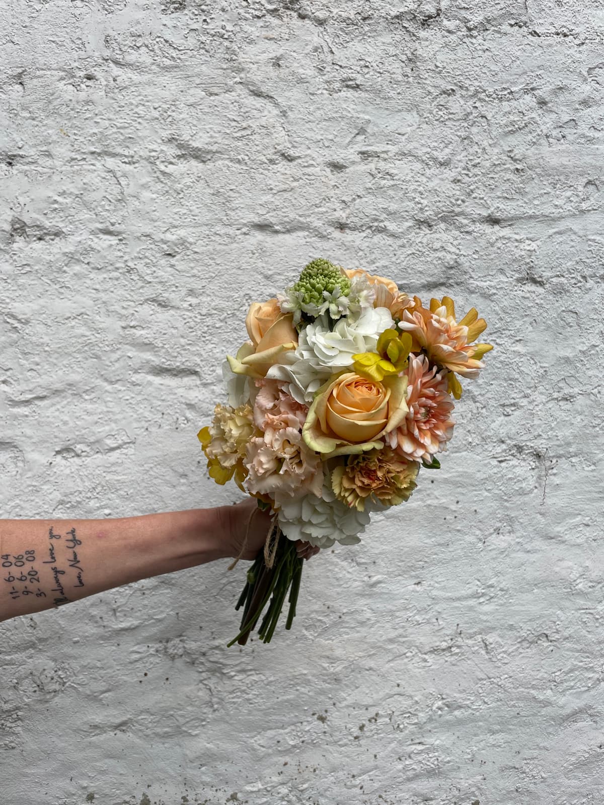 A tattooed arm holds a lush, hand-tied floral bouquet against a textured white brick wall. The arrangement features peach roses, apricot dahlias, creamy hydrangeas, and pale yellow carnations tied with simple twine.