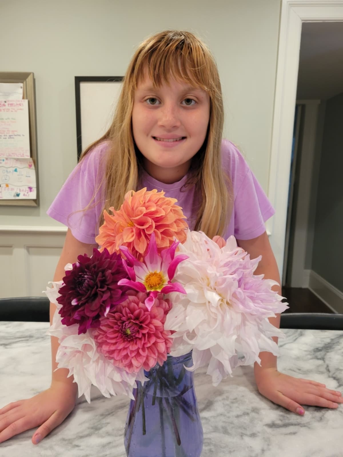A smiling young girl stands behind a blue glass vase filled with a colorful assortment of dahlias on a marble countertop. The bouquet features large pale pink blooms, vibrant coral flowers, and deep burgundy accents.