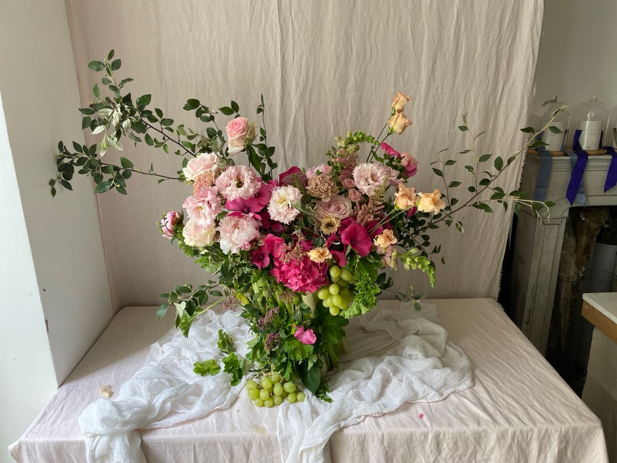 A lush, asymmetrical floral arrangement featuring blush lisianthus, magenta clematis, and clusters of green grapes, set against a neutral draped background.