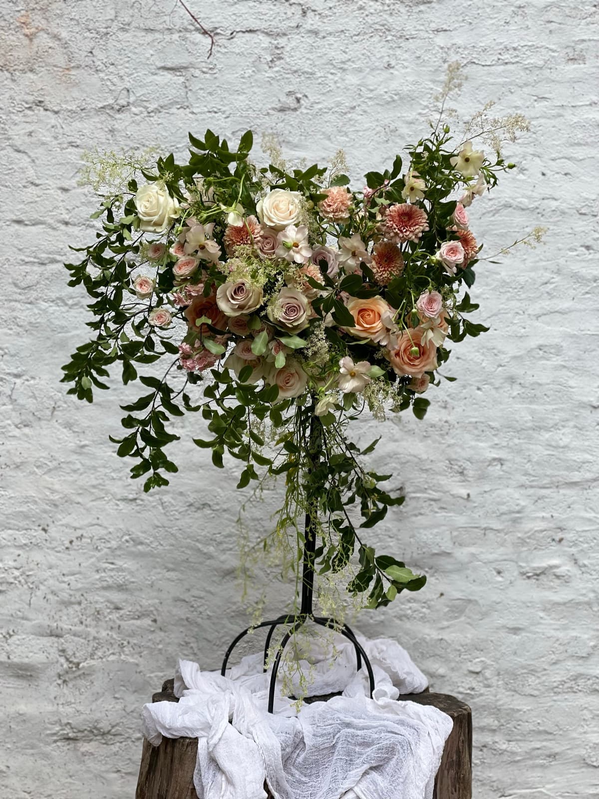 A lush, heart-shaped floral arrangement on a tall black metal stand against a white brick wall. The design features dusty mauve and apricot roses, small pink chrysanthemums, and white Queen Anne's lace mixed with trailing dark green vines. The stand rests on a wooden stump covered in white cheesecloth.