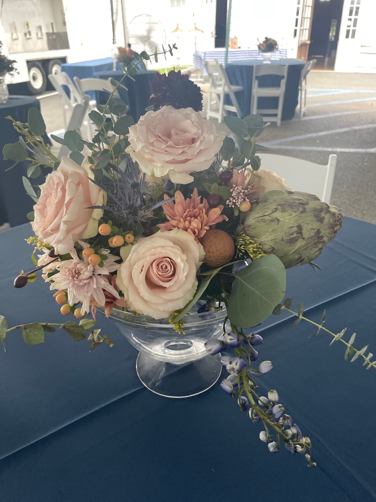 A lush floral centerpiece in a glass compote featuring blush roses, a large green artichoke, blue thistle, purple lupine, and peach berries on a blue tablecloth.