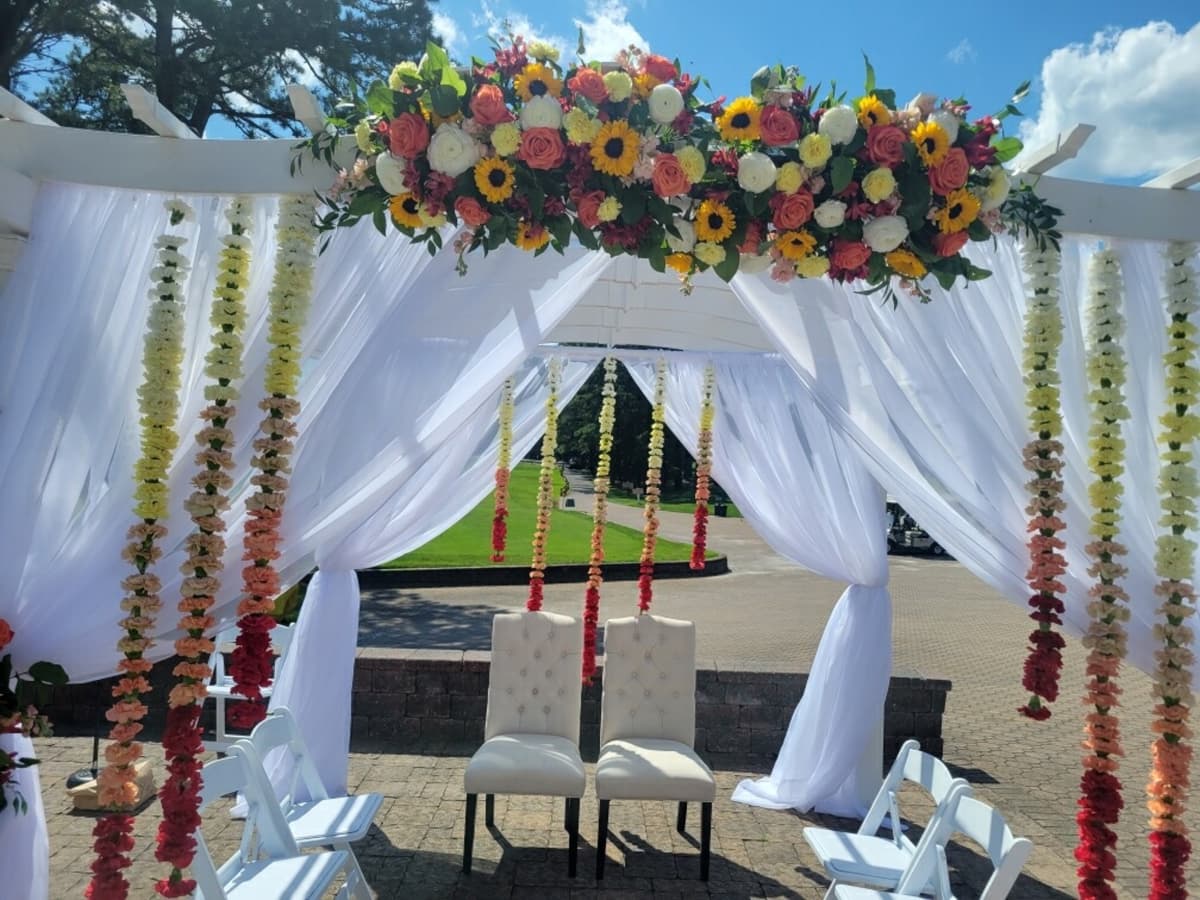 Outdoor wedding ceremony structure draped in white fabric, featuring a top arrangement of sunflowers and roses, with hanging ombre floral garlands transitioning from white to deep red behind two beige chairs.