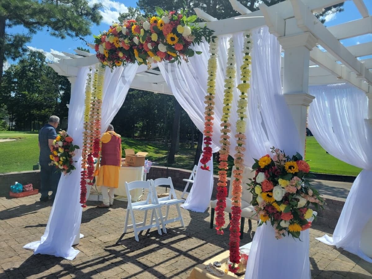 A white outdoor wedding pergola draped in sheer white fabric, decorated with a top spray of sunflowers and roses, and featuring hanging vertical garlands of carnations in an ombré gradient from yellow to red.