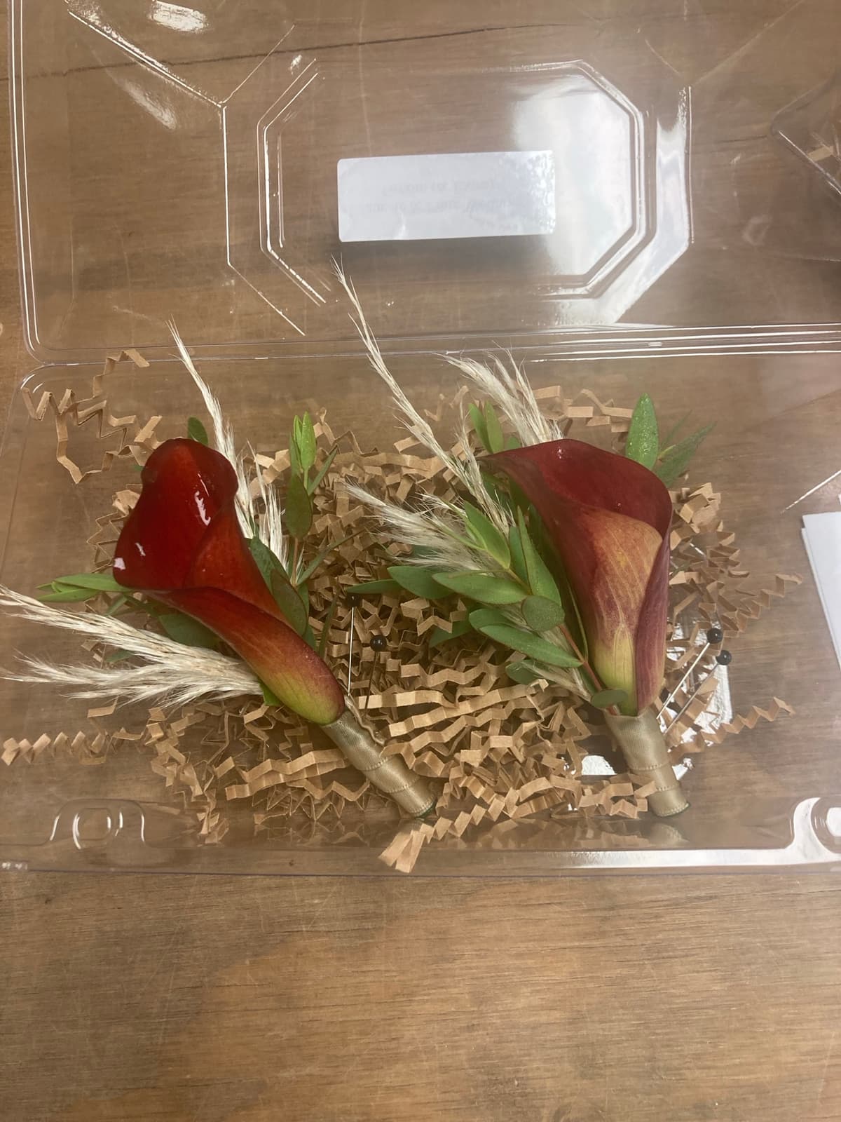 Two burgundy calla lily boutonnieres with dried grass accents resting on crinkle paper in a clear box.