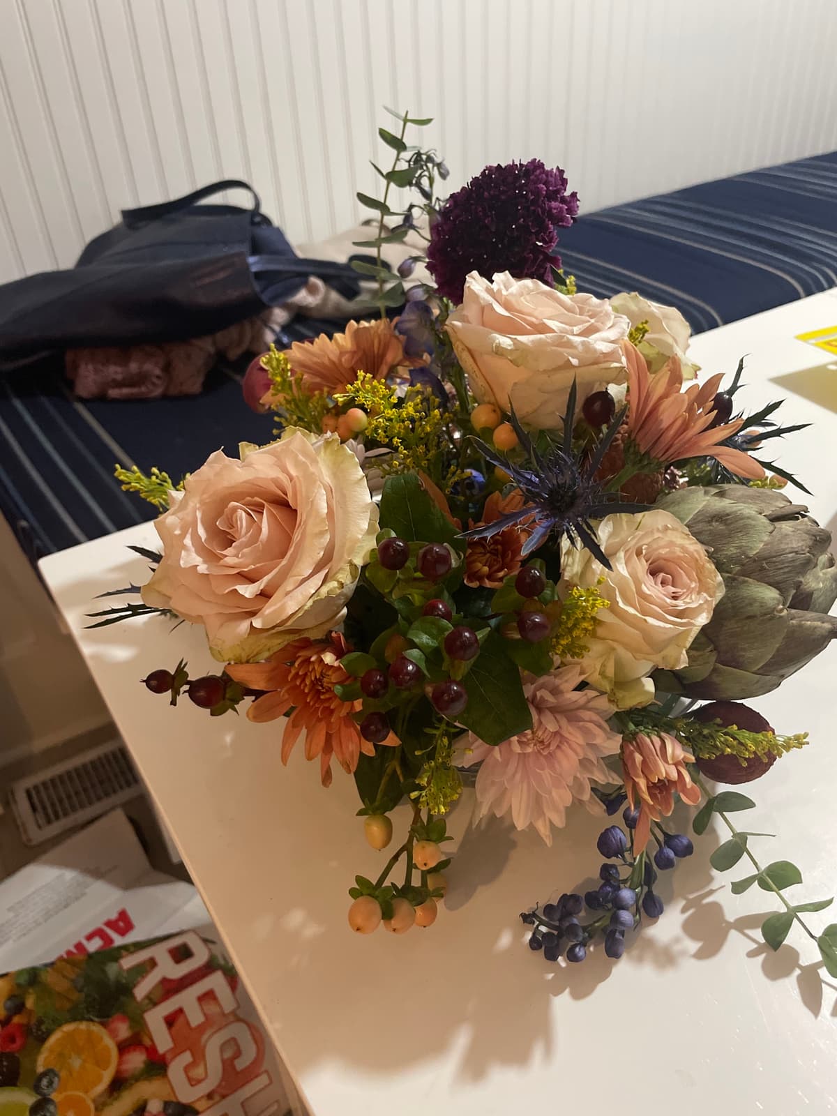 A lush, textural floral arrangement featuring a large green artichoke, blush roses, peach chrysanthemums, blue thistle, and deep red berries on a white table.