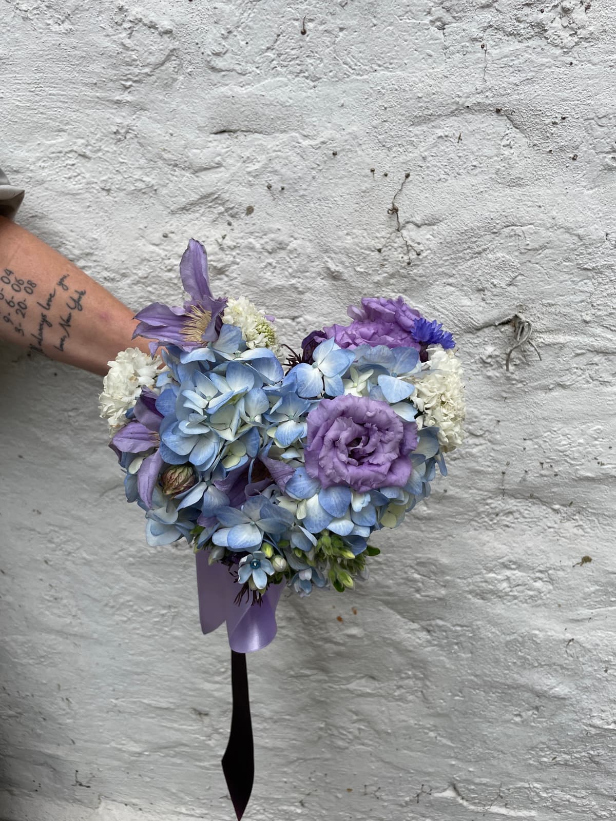 A hand holding a lush bouquet of blue hydrangeas, purple lisianthus, and white scabiosa against a textured white wall, tied with a purple ribbon.