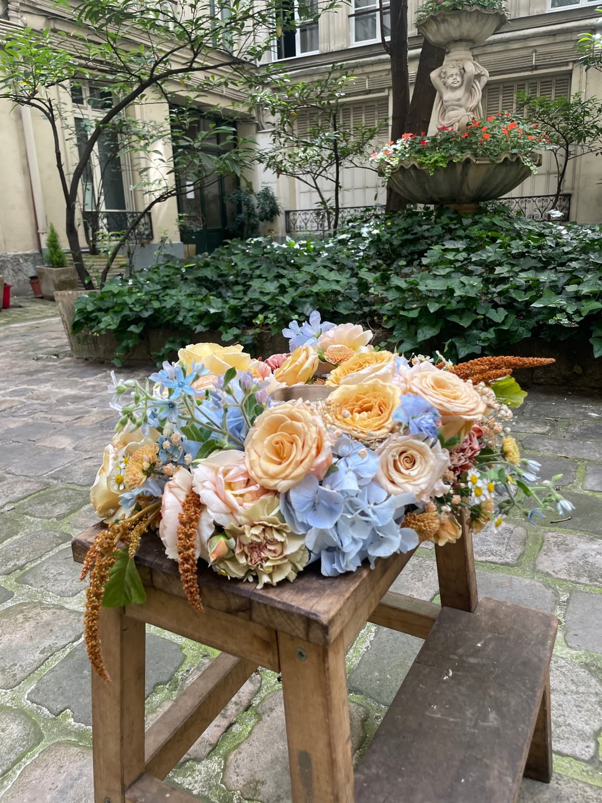 A lush floral arrangement sitting on a wooden stool in a stone courtyard, featuring peach roses, blue hydrangeas, and bronze amaranthus.