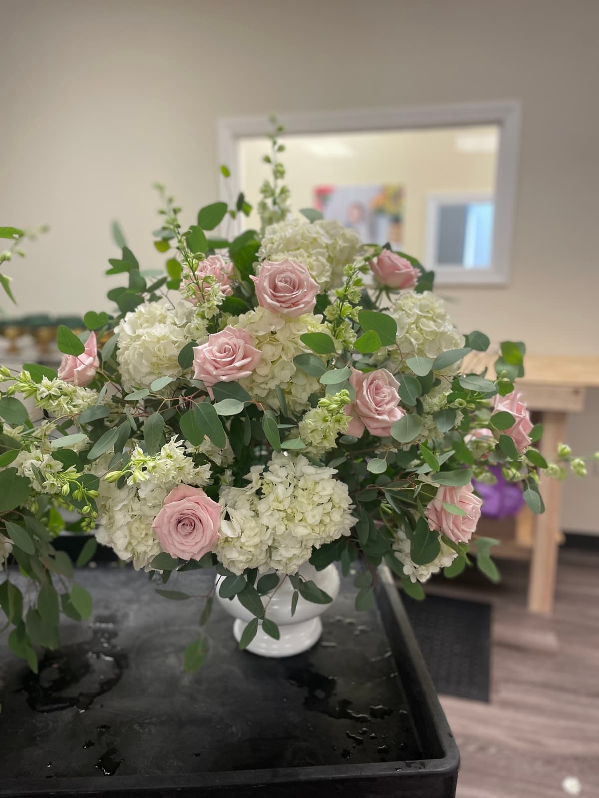 A lush floral arrangement featuring soft pink roses, white hydrangeas, and white larkspur mixed with eucalyptus greenery in a white ceramic urn vase.