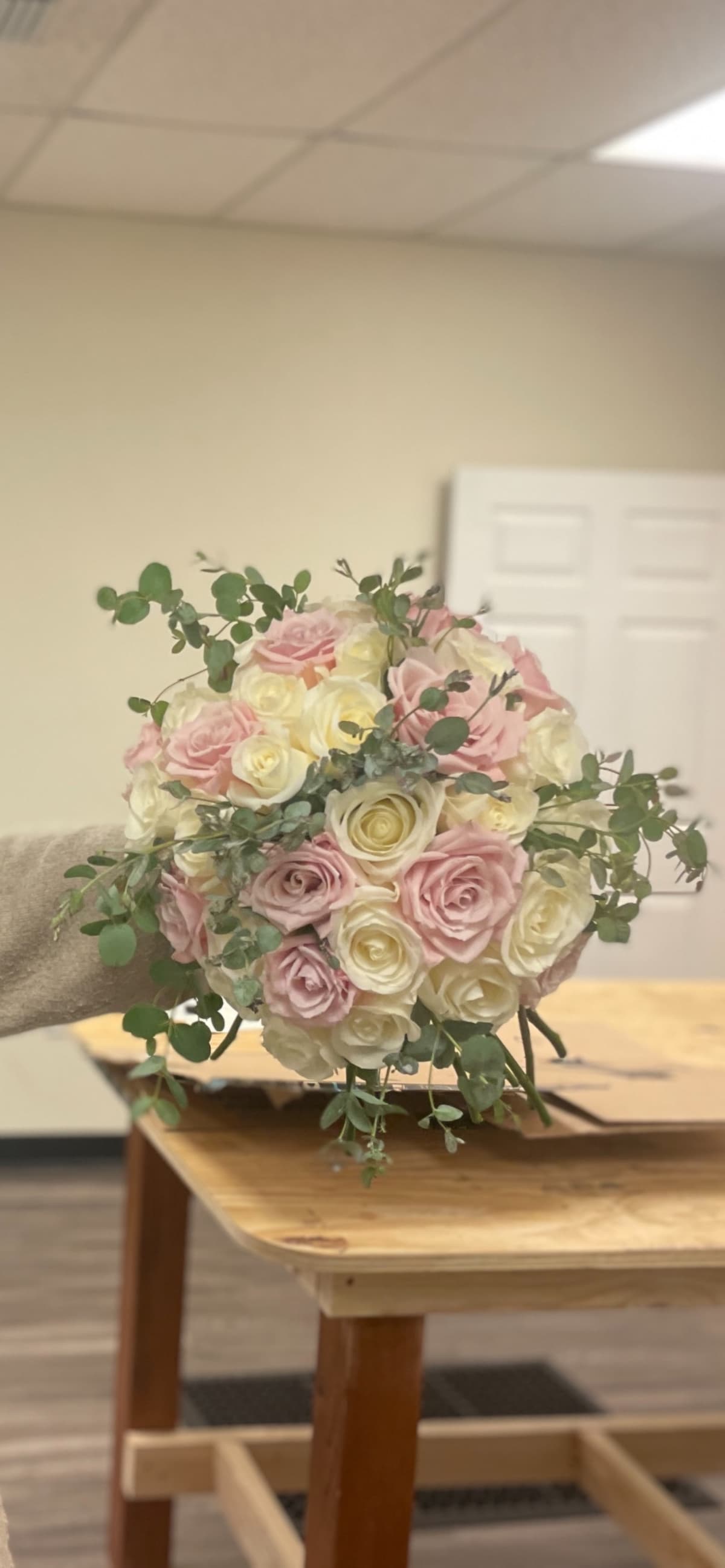 A round, hand-held bouquet featuring blush pink and creamy white roses mixed with sprigs of green eucalyptus leaves, held over a wooden table in a floral studio.