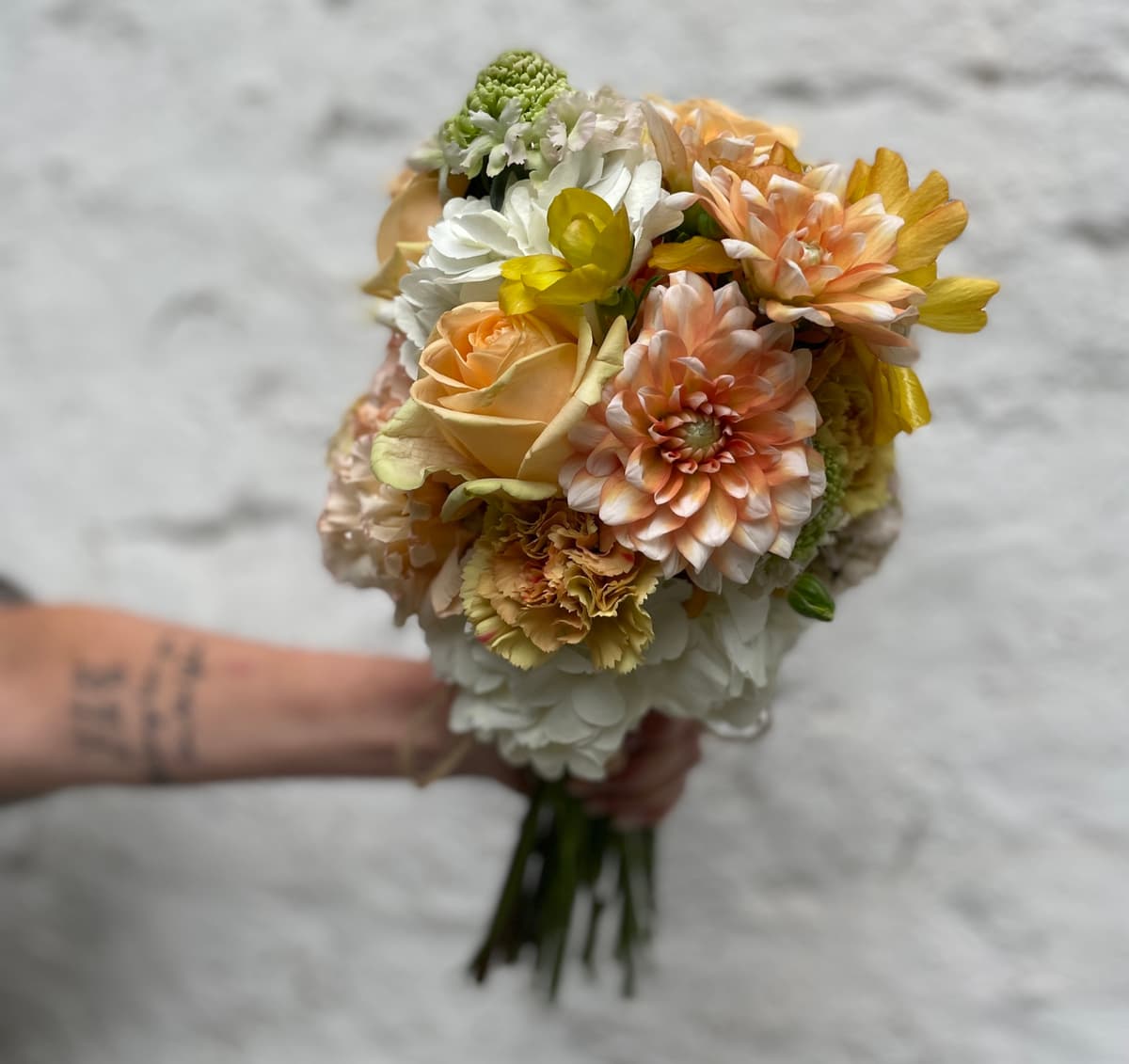 A person holding a lush, round bouquet of flowers against a white textured wall. The arrangement features peach dahlias, creamy roses, yellow freesias, and ruffled carnations.