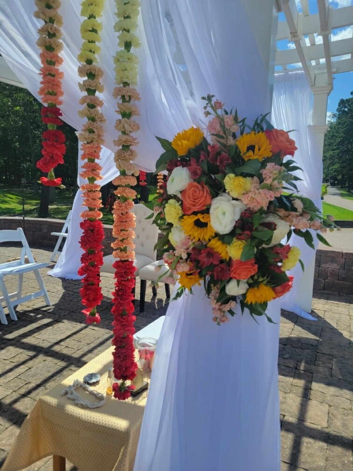 Outdoor wedding ceremony structure draped in white fabric featuring hanging ombre carnation garlands transitioning from yellow to red, alongside a large floral pillar spray with sunflowers, roses, and ranunculus.