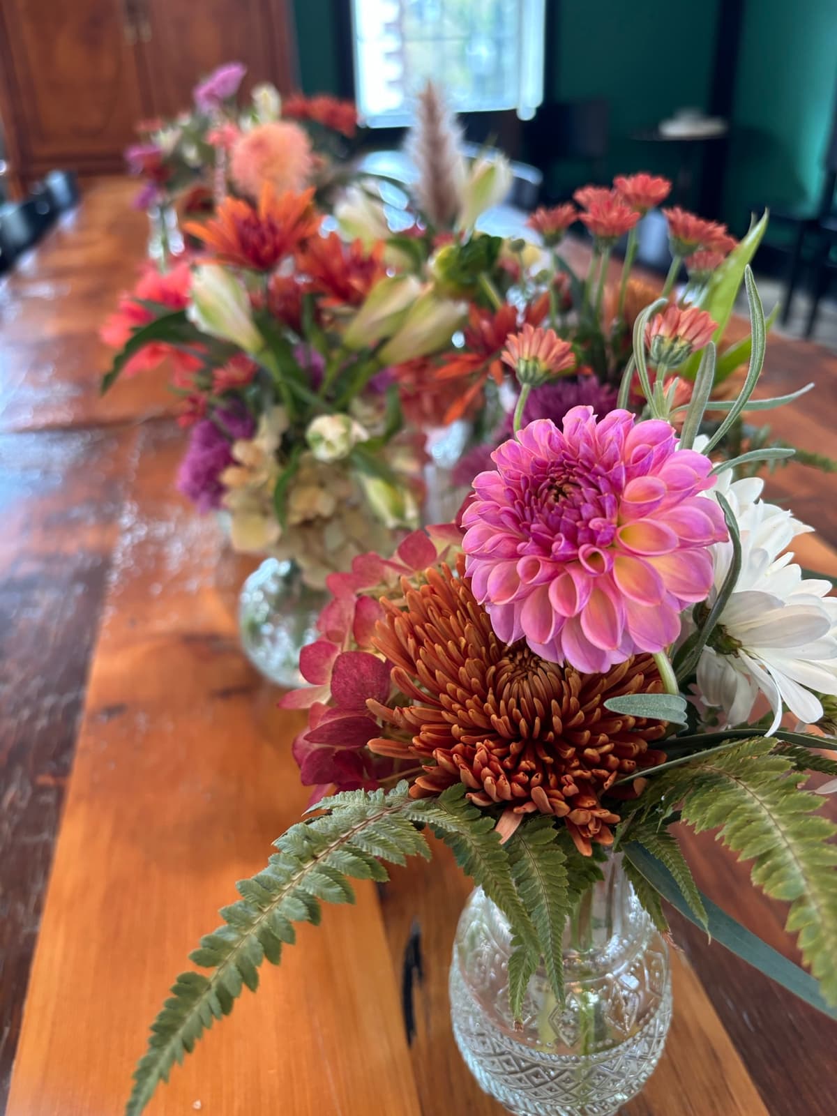 Close-up of a floral arrangement in a cut-glass vase on a wooden table, featuring a bright pink dahlia, a rust-colored spider mum, white blooms, and green fern leaves.