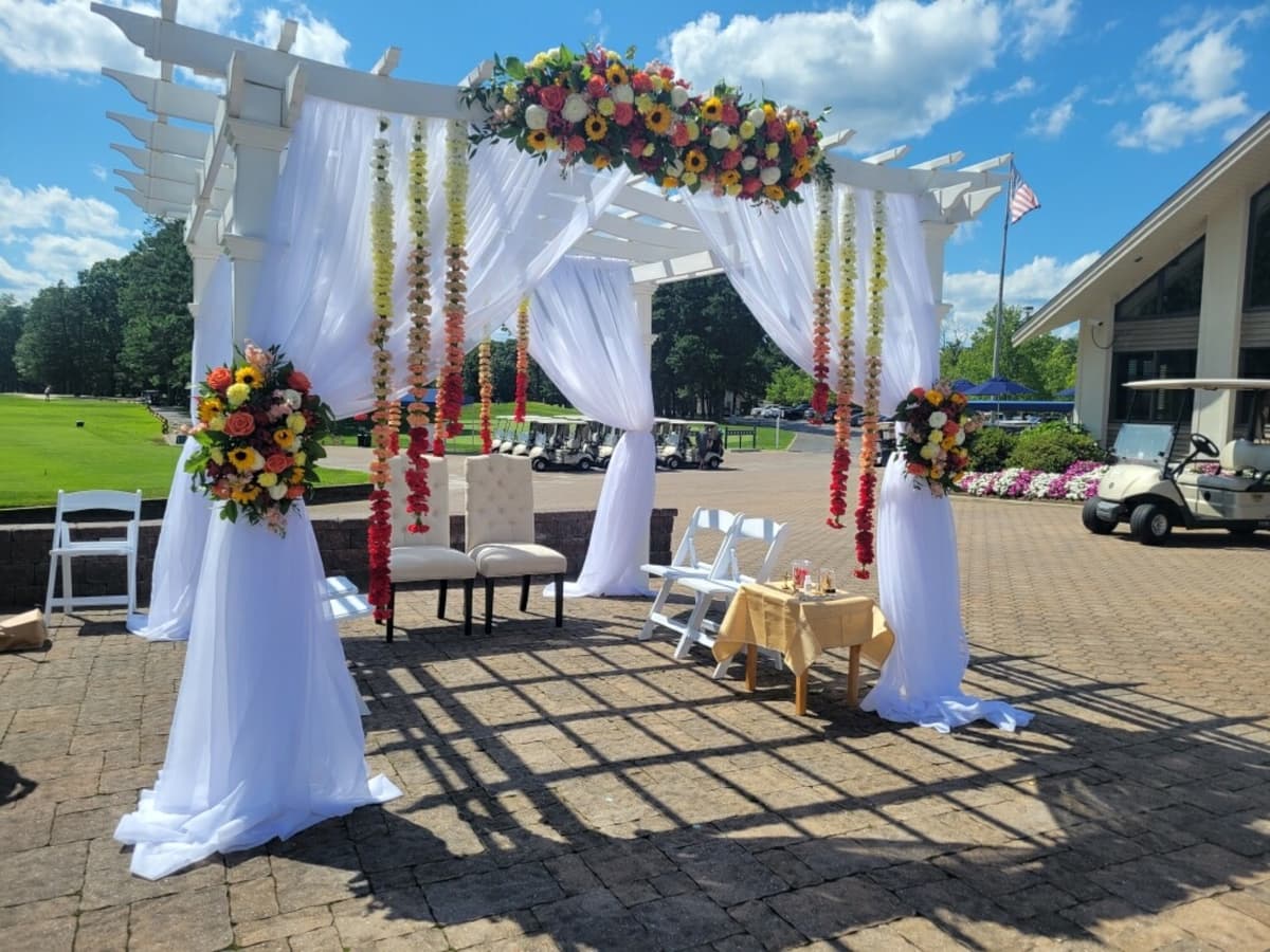 Outdoor wedding ceremony setup under a white pergola with sheer white draping. Vertical floral garlands in ombre shades of yellow to red hang from the beams. Large floral arrangements featuring sunflowers and coral roses adorn the top and sides. Two beige chairs sit beneath the structure on a paved patio.