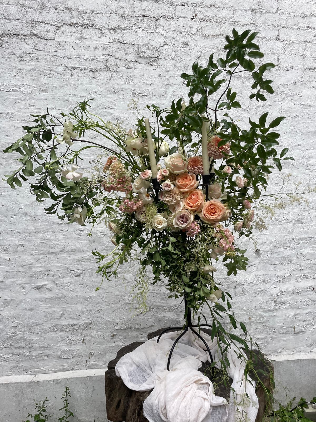 A lush, asymmetrical floral arrangement featuring peach and dusty lavender roses, small pink dahlias, and airy Queen Anne's lace. Long, wild branches of greenery extend outward against a white brick wall. The arrangement sits on a rustic wooden base draped with white cheesecloth fabric.