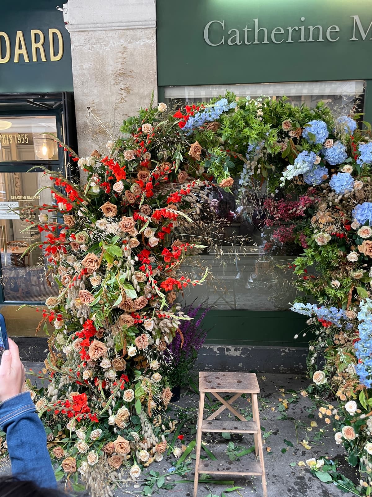 A lush, asymmetrical floral arch framing a green storefront window. The arrangement features blue hydrangeas, sandy beige roses, and bright red gladiolus mixed with dried grasses and greenery. A wooden ladder stands to the right, suggesting an installation in progress.