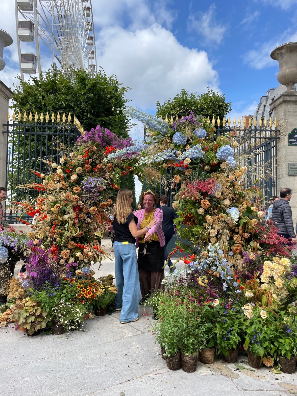 A massive, lush floral installation covering an iron gate outdoors, featuring a mix of fresh and dried elements. The arrangement includes peach and antique-colored roses, blue hydrangeas, purple delphiniums, and dried grasses. Two women are standing in front of the archway, and a Ferris wheel is visible in the background against a blue sky.