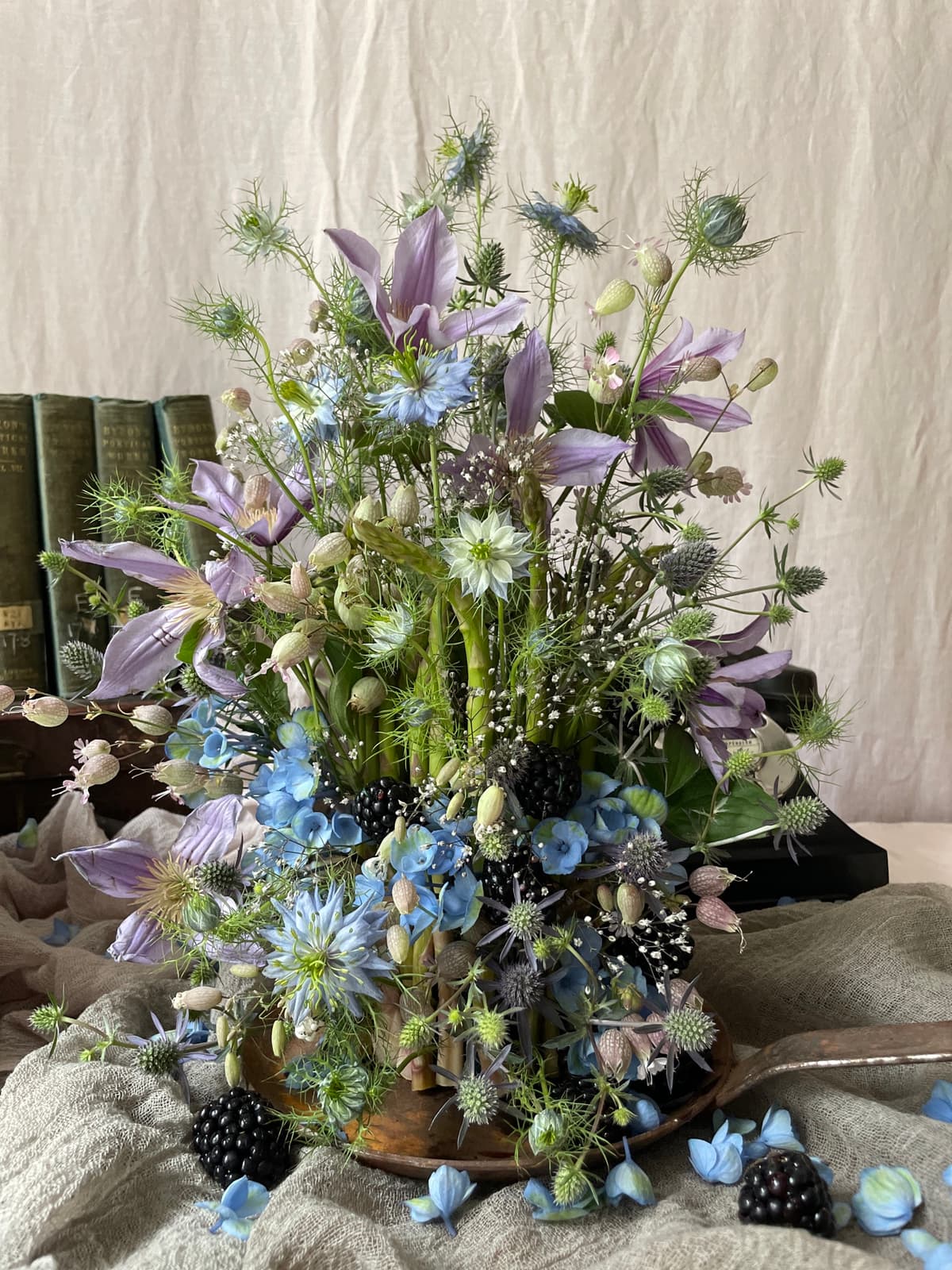 A wild, garden-style floral arrangement featuring lavender clematis, blue nigella, and dark blackberries arranged on top of vintage books against a linen background.